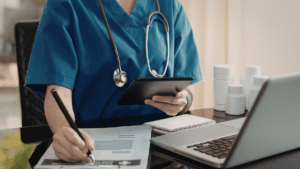 Healthcare professional completing payroll and timesheet administration on a laptop and tablet in a clinical setting.