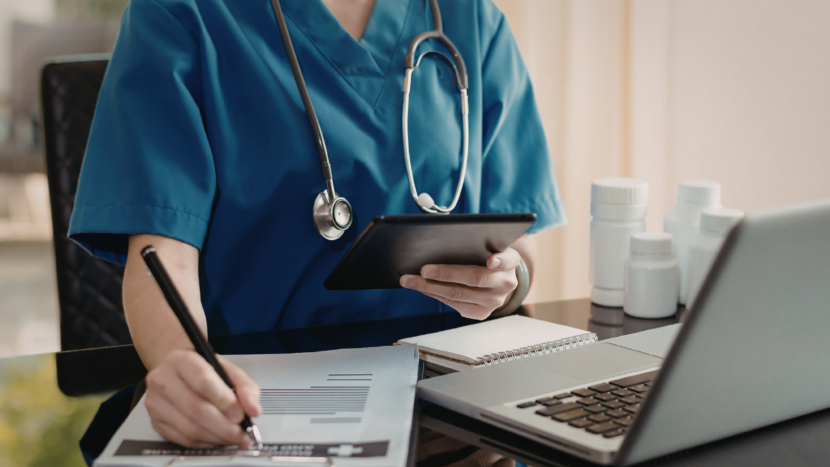 Healthcare professional completing payroll and timesheet administration on a laptop and tablet in a clinical setting.